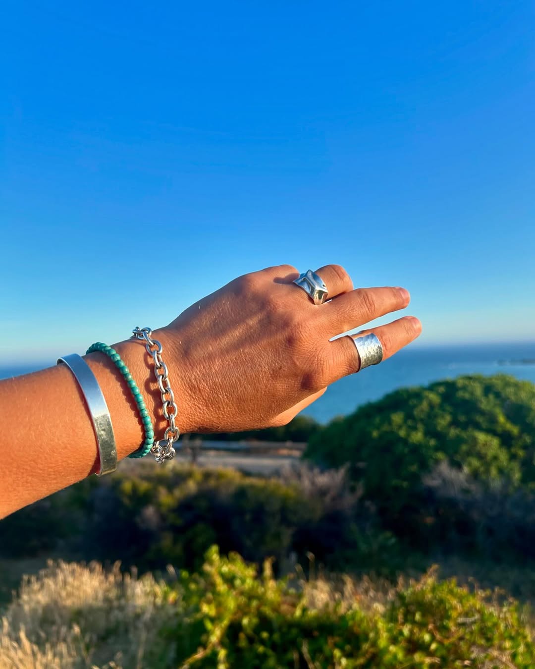 A hand stacked with a silver cuff, turquoise beads, a link bracelet and two silver rings, held over a coastal headland.