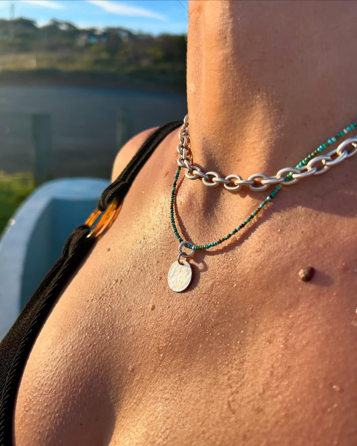 An oval-link silver chain and a turquoise bead strand with a small hammered pendant, worn in golden hour light.